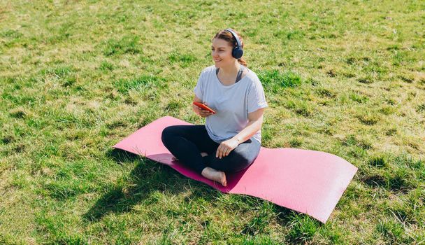 Красивая молодая рыжая женщина расслабляется со смартфоном на коврике для йоги после тренировки. Счастливая девушка с наушниками прослушивания музыки или просмотра Интернета. Концепция спорта или йоги - Фото, изображение