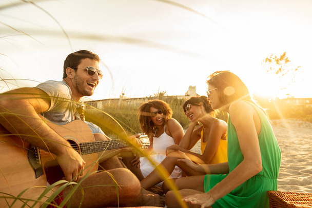 Счастливая карферная жизнь молодых друзей, наслаждающихся свободой летних каникул, сидя за гитарой на пляже на закате - Фото, изображение