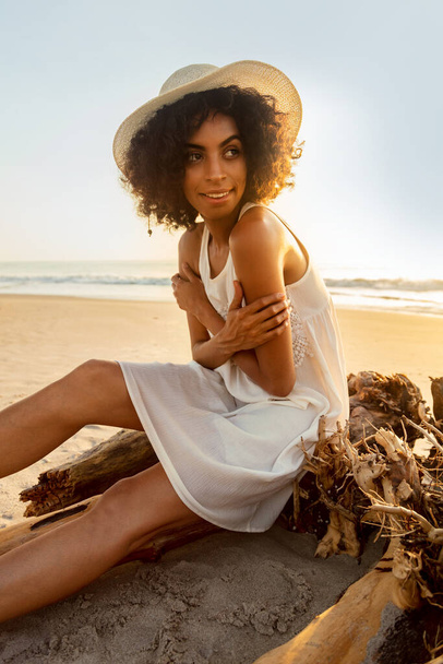 Sonriente mujer afroamericana en sombrero en la playa al amanecer sentada en madera a la deriva disfrutando de vacaciones de verano en solitario sin preocupaciones - Foto, Imagen