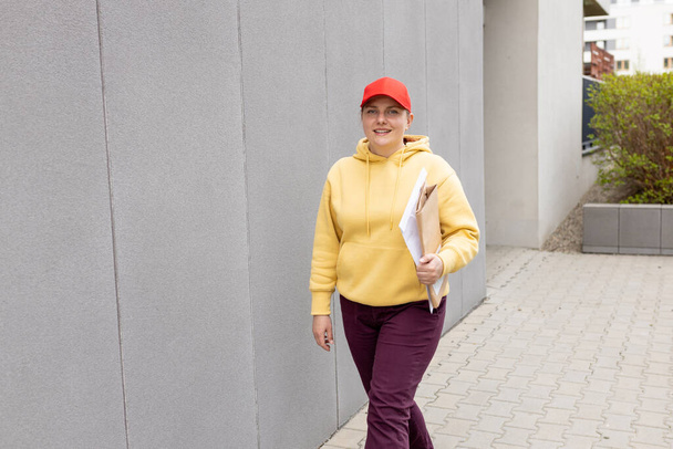 Toimitus nainen punainen lippis odottaa asiakasta lähellä rakennuksen moderni kaupunki, Kuriiri Toimituspalvelu. Nainen punaisessa hatussa laatikoilla sisäänkäynnin lähellä - Valokuva, kuva