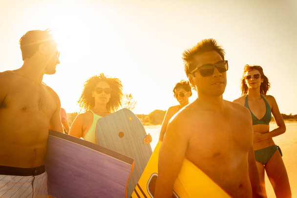 Outdoor fun on beach at sunset with multi ethnic friends in swimwear carrying bodyboards enjoying Summer vacation together - Φωτογραφία, εικόνα