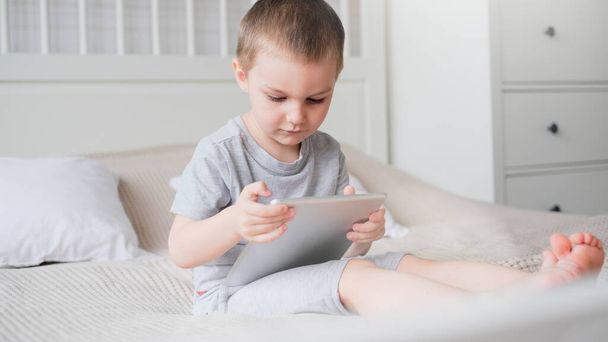 Un pequeño chico caucásico en una habitación de luz con una camiseta gris juega con una tableta mientras está sentado en el sofá.  - Foto, Imagen