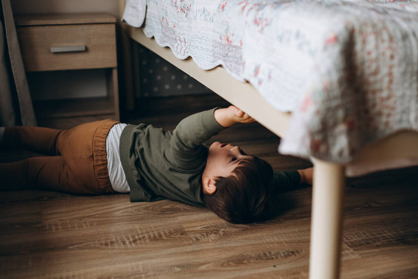 Little, pensive, two years old boy sitting under the bed and playing with toy children's car.  - Fotó, kép