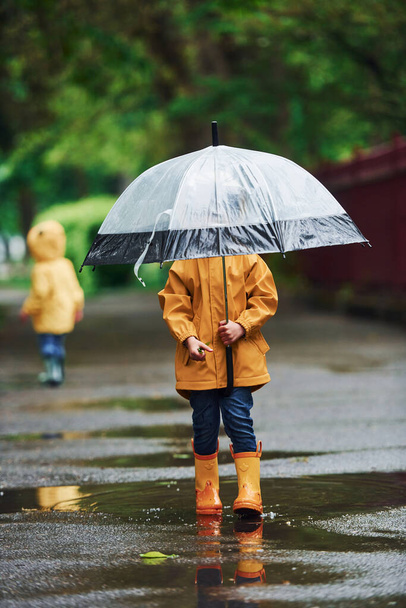 Two kids with umbrella in yellow waterproof cloaks and boots playing outdoors after the rain together. - Photo, Image