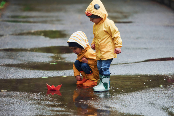 Two kids in yellow waterproof cloaks and boots playing with handmade paper boat outdoors after the rain together. - Photo, image