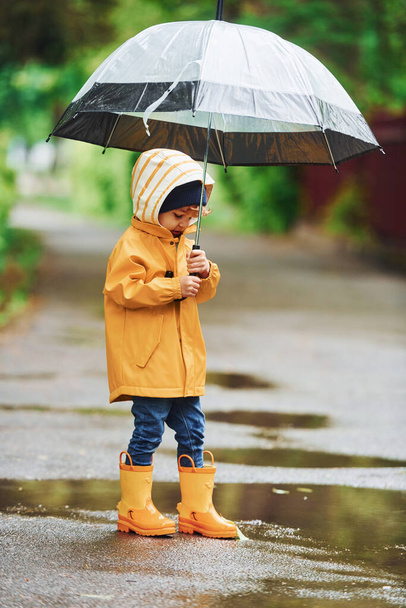 Kid in yellow waterproof cloak, boots and with umbrella playing outdoors after the rain. - Foto, Imagen