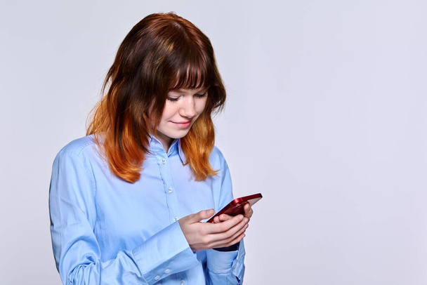 Smiling teenage female looking at smartphone screen, on light studio background, copy space - 写真・画像