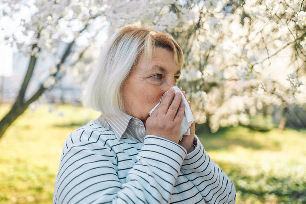 50s woman sneezing in park. Allergy, flu, virus concept. - 写真・画像
