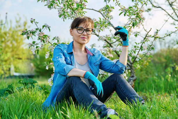 Positive beautiful mature female gardener sitting on the grass with blossoming apple tree. - 写真・画像