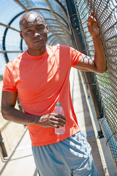 Τοποθετήστε νέους Afro Αμερικανός άνθρωπος ενυδάτωση με νερό κατά τη διάρκεια της προπόνηση και στηρίζεται κατά σύρμα φράχτη σε καλυμμένο διάδρομο - Φωτογραφία, εικόνα