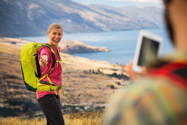 Happy Caucasian man met behulp van smart phone technologie het nemen van een reis foto van zijn vrolijke glimlachende vrouw wandelen The Remarkables Queenstown Nieuw-Zeeland - Foto, afbeelding