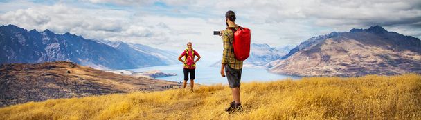 Panoramic backpacking male using smartphone technology taking travel pictures of his girlfriend achieving her goals hiking in The Remarkables Queenstown New Zealand - Photo, Image
