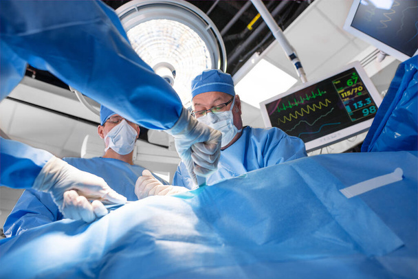 Caucasian surgical team wearing scrubs with face masks and gloves operating on patient with overhead light and monitor - Photo, Image