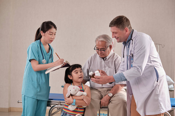 Professional Caucasian male doctor advises grandfather and grandchild, explains prescription with medical trainees at patient hospital bed, illness consultations, and family health examination clinic. - Foto, Imagem