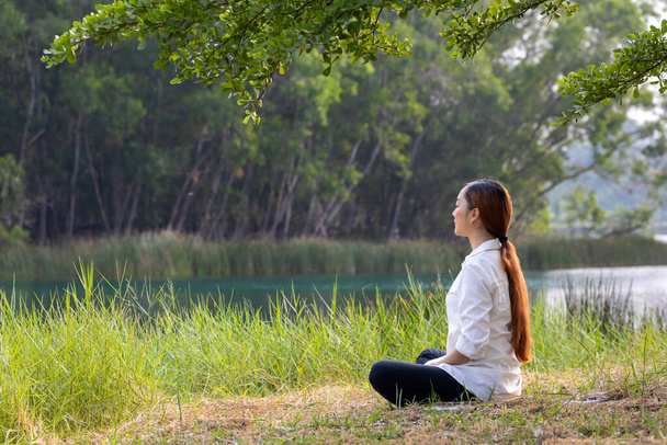 Mujer sentada relajadamente y practicando meditación en el parque público para alcanzar la felicidad desde la paz interior sabiduría bajo el árbol durante el verano - Foto, Imagen
