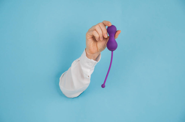 Female hand sticking out of a hole in a blue background and holding a kegel machine.  - Foto, Imagen