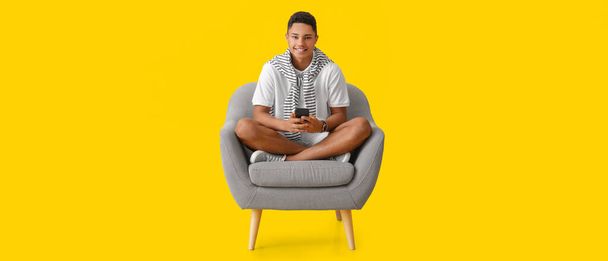 Young African-American guy with mobile phone sitting in armchair on yellow background - Zdjęcie, obraz