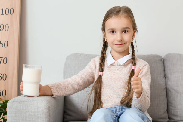 klein meisje met glas melk zitten in fauteuil en tonen duim omhoog - Foto, afbeelding