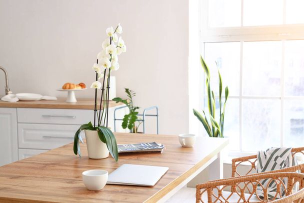 Красивый цветок орхидеи, чашка и ноутбук на обеденном столе в светлой кухне - Фото, изображение
