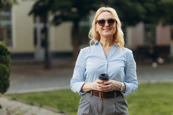 Πορτρέτο μιας υψηλόβαθμης επιχειρηματία που κρατάει ένα χάρτινο κύπελλο. Ηλικιωμένη χαμογελαστή ευτυχισμένη γυναίκα στα 50 της περπατά στην πόλη. Business Concept επιτυχία εργασίας - Φωτογραφία, εικόνα