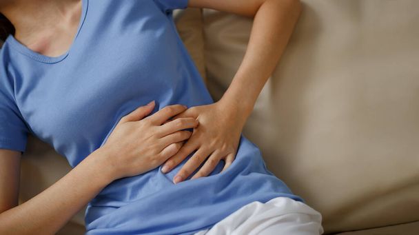 Woman having stomach aches hard, suffering from abdominal pain while sitting on sofa at home. - Valokuva, kuva