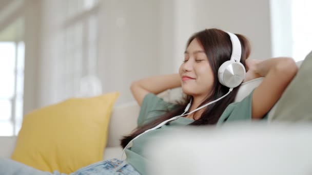 Genç Asyalı kadın evdeki salonda kanepede müzik dinliyor. Mutlu Asyalı kadın cep telefonu kullanıyor, kulaklık takıyor ve kanepede oturuyor. - Video, Çekim