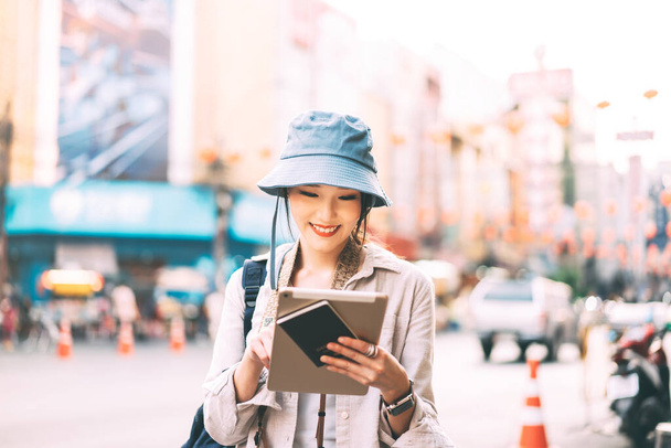Atraente feliz jovem adulto asiático mulher viajante usar chapéu azul e mochila. Solo viajando com ensino moderno usando tablet para procurar estilo de vida na cidade ao ar livre no dia. Conceito de viagem de verão. - Foto, Imagem