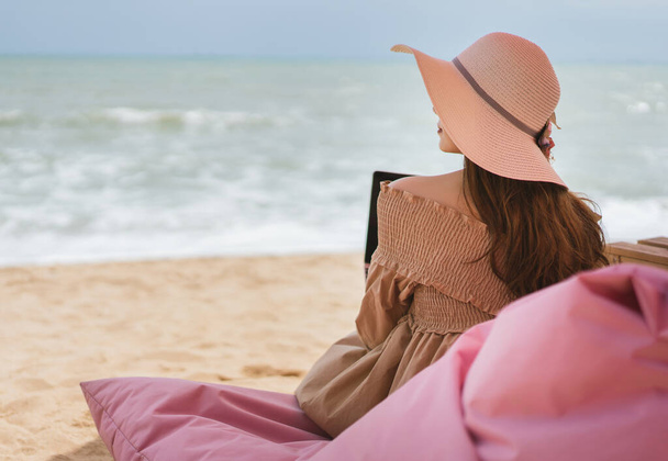 On vacation at Pattaya beach, asian traveler woman with floppy hat relax from working with laptop on a pink chair in luxury cafe. - Фото, зображення