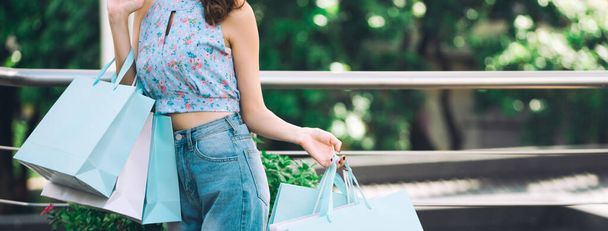 Shopper asian woman holding shopping bags. Her face with happy smile and wear blue shirt. Customer enjoy summer sale city lifestyle concept. Banner size background. - 写真・画像