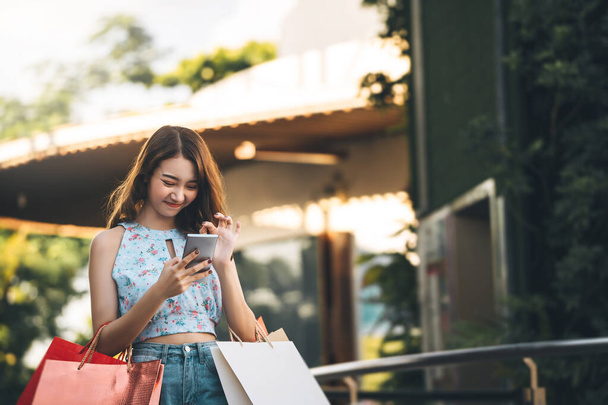 Wohlbefinden Single-Lebensstil im Urlaub Junge erwachsene asiatische Frau nutzt Handy für Online-Anwendung und hält Einkaufstüten.  - Foto, Bild