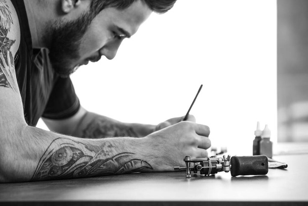 Professional tattoo artist drawing sketch at table indoors. Black and white photography - Φωτογραφία, εικόνα