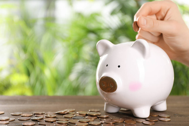 Woman putting coin into piggy bank at wooden table against blurred green background, closeup. Space for text - Foto, afbeelding