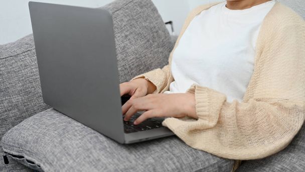 Urocza azjatycka młoda kobieta relaksuje się na kanapie w salonie i przy użyciu laptopa. Praca zdalna w domu - Zdjęcie, obraz