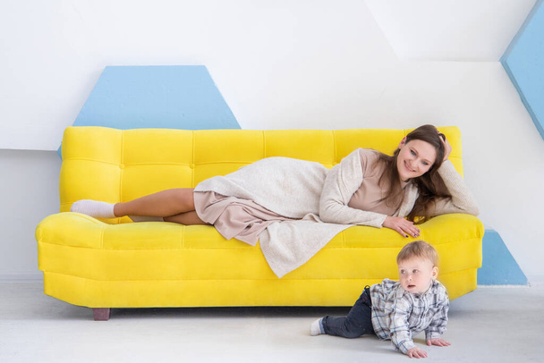 Jeune femme caucasienne heureuse se trouve sur un canapé jaune et regarde son bébé ramper sur le sol. Attendre les premiers pas du bébé. - Photo, image