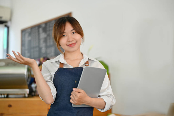 Mooie charmante Aziatische jonge vrouwelijke coffeeshop ondernemer of barista staan in de coffeeshop en holding tablet. - Foto, afbeelding