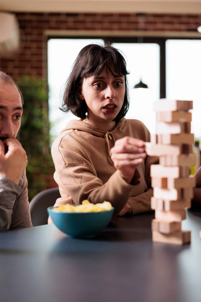 Worried woman carefully removing wood block from wooden tower structure while sitting at table. - Photo, image
