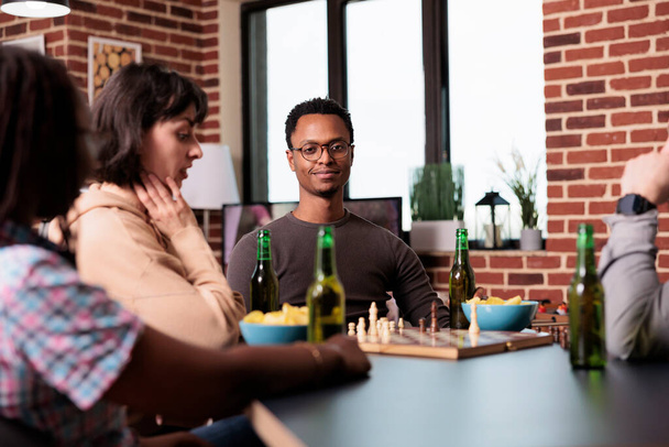 Hombre sonriente sentado en la mesa en la sala de estar con amigos mientras ve una partida de ajedrez. - Foto, Imagen