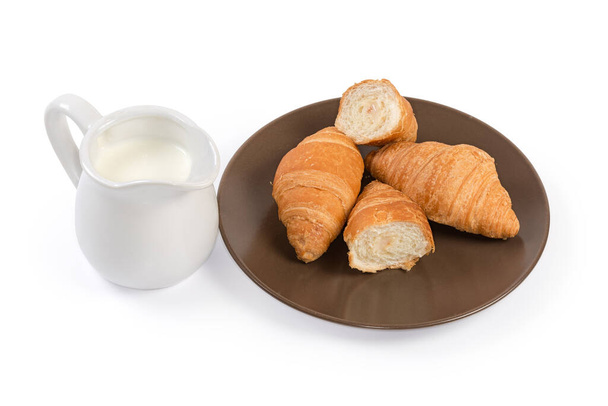 Celé a půlky malých croissantů z listového těsta s kondenzovanou mléčnou náplní na hnědém talíři, mléko v malém džbánu na bílém pozadí - Fotografie, Obrázek