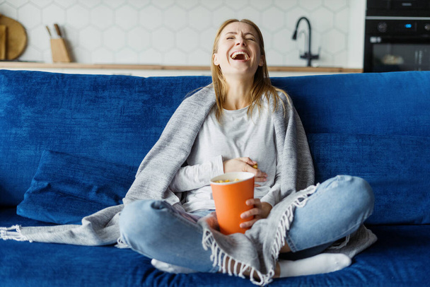 Happy woman laughing loud while she sitting on sofa with popcorn and watching comedy movie or hilarious video on TV. Female spend weekend at home, having fun, eating snacks, look entertainment program - Photo, image
