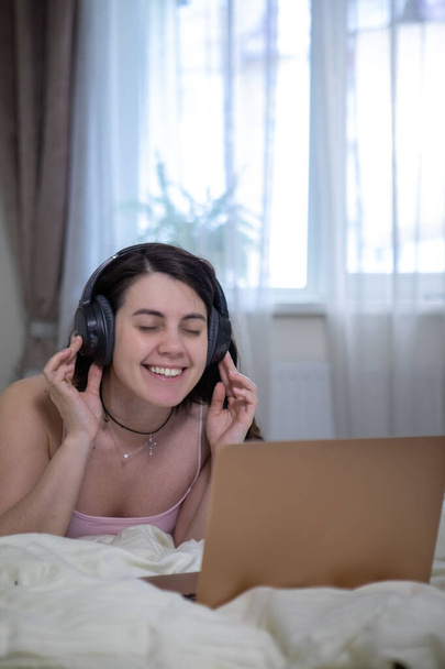 Frau legt sich ins Bett, arbeitet am Laptop und hört Musik im Kopfhörer - Foto, Bild