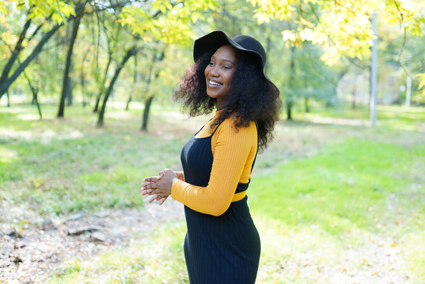 Hermosa mujer afroamericana sonriendo y caminando en el parque durante el atardecer. Retrato al aire libre de una chica negra sonriente - Foto, Imagen