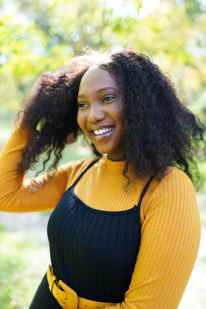 Όμορφη Αφροαμερικανή γυναίκα χαμογελά και περπατάει στο πάρκο κατά τη διάρκεια του ηλιοβασιλέματος. Εξωτερική πορτρέτο ενός χαμογελαστού μαύρου κοριτσιού - Φωτογραφία, εικόνα