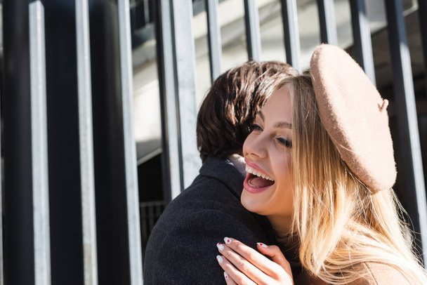 excited blonde woman in beret laughing near boyfriend outdoors - Fotó, kép