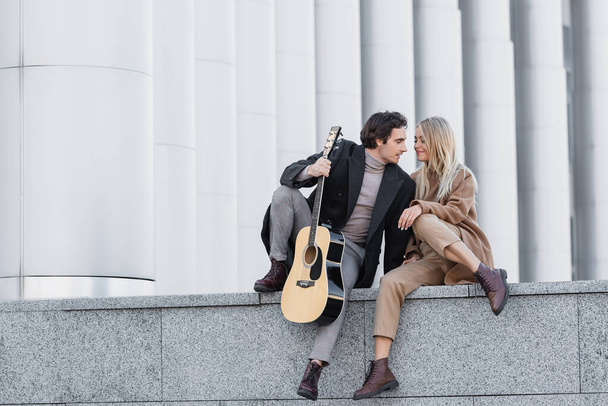 Sonbahar kıyafetli bir kadın korkulukta gitar çalan bir adamın yanında oturuyor. - Fotoğraf, Görsel