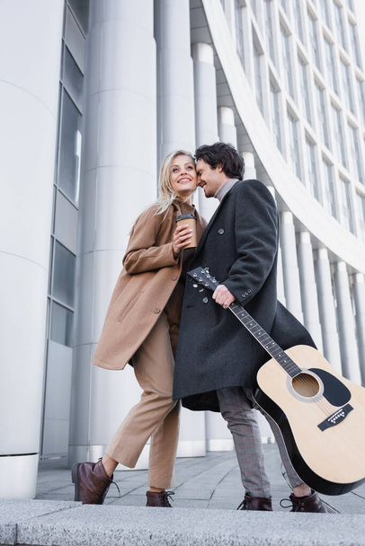 アコースティックギターとコーヒーのコートを着たカップルがビルの近くに立っているのを見ると - 写真・画像