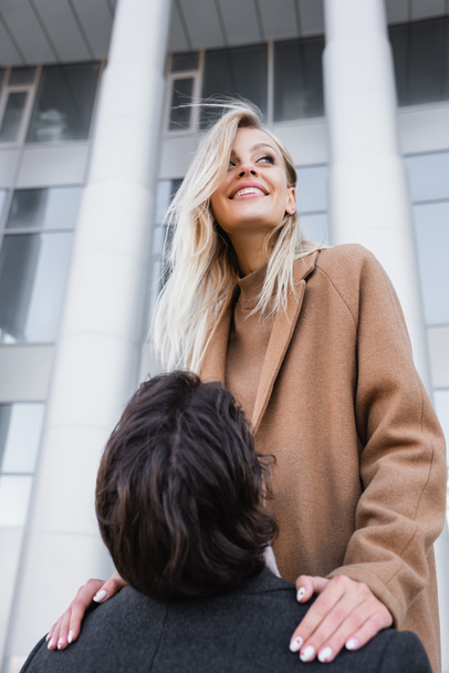 man raising up cheerful blonde woman in coat on urban street - Photo, image