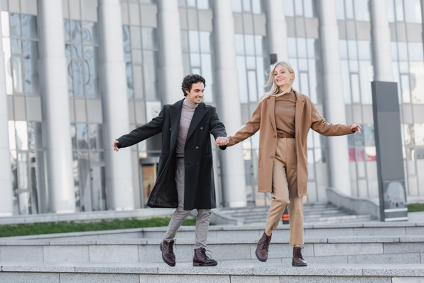 full length of excited couple in coats and leather boots holding hands and running on street - Fotografie, Obrázek