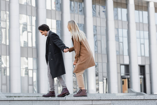 side view of excited couple in leather boots and coats holding hands and walking on parapet - Photo, Image