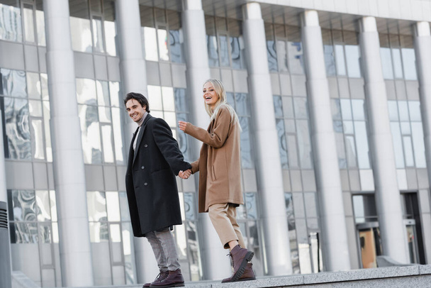 full length of cheerful couple in leather boots and autumn coats holding hands near blurred building - Foto, Bild
