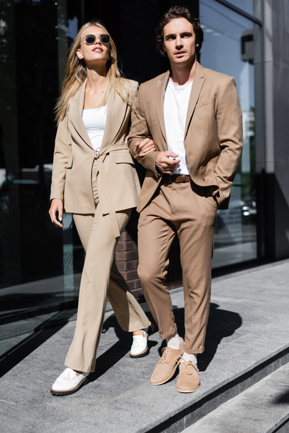 full length of stylish couple in beige suits walking on urban street - Фото, изображение